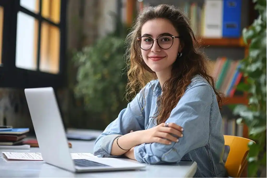 Confident young woman wearing glasses and smiling for the camera. She's on a library, working on a laptop. The image illustrates how online tutoring is a great side hustle to earn an online income.